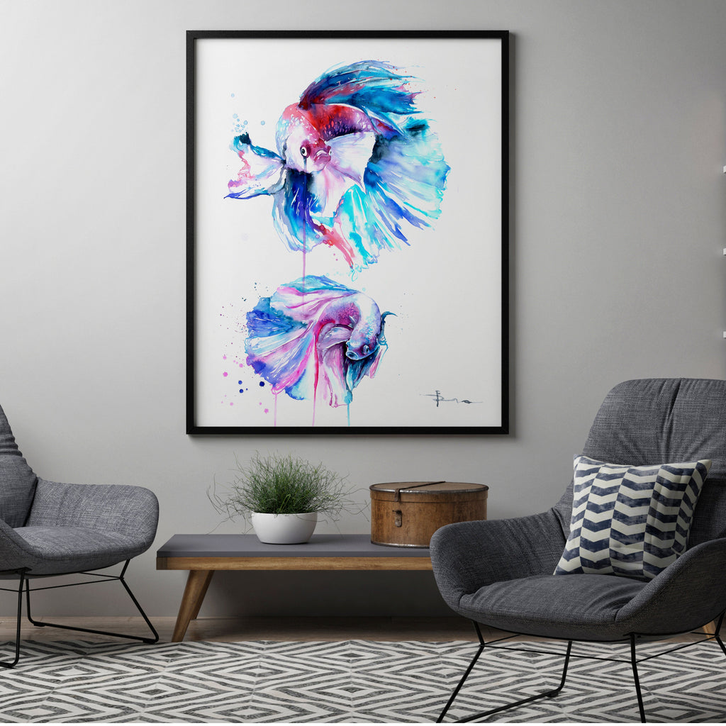 A framed watercolor art print featuring two colorful fish, displayed in a modern living room setting between two gray armchairs and a bookshelf.