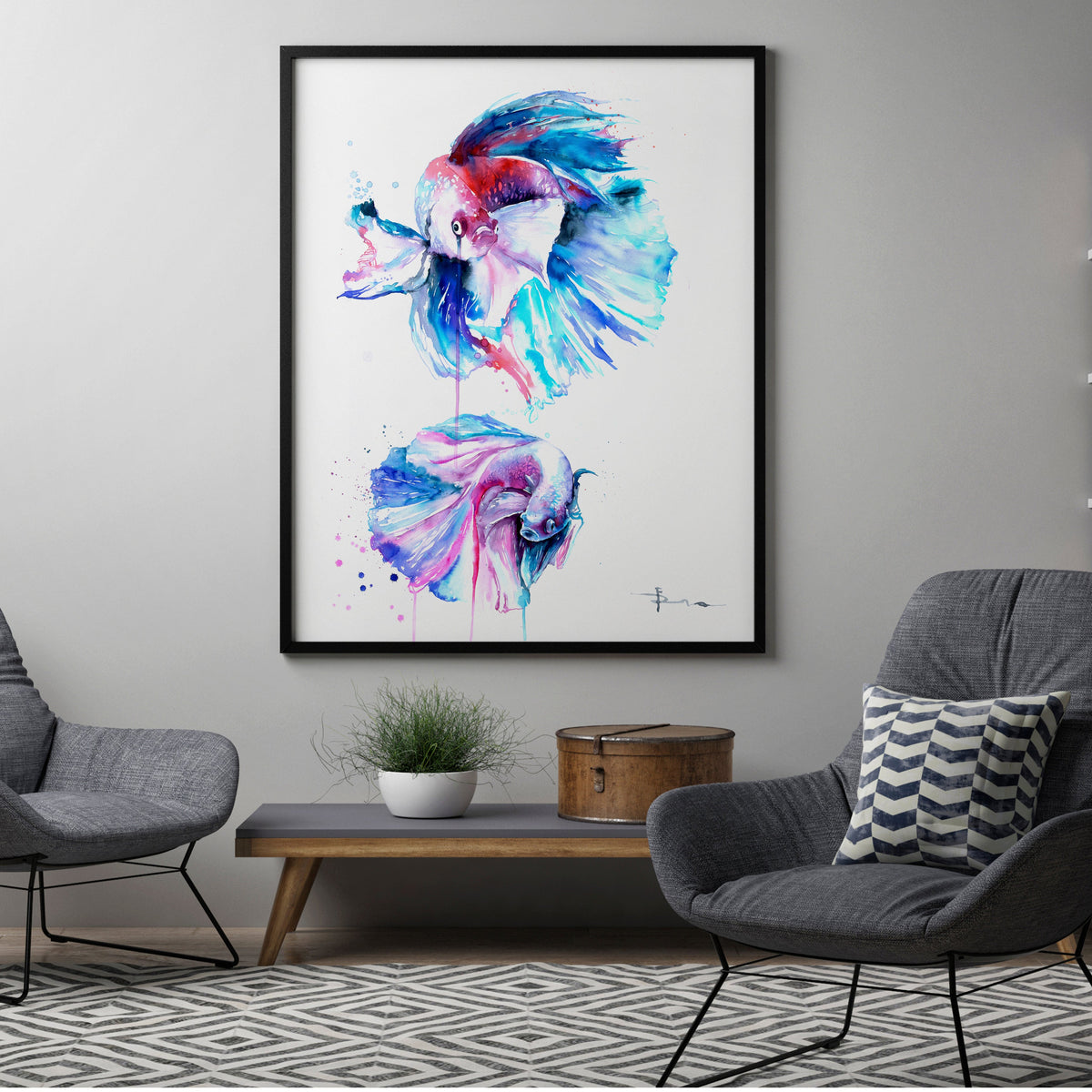 A framed watercolor art print featuring two colorful fish, displayed in a modern living room setting between two gray armchairs and a bookshelf.